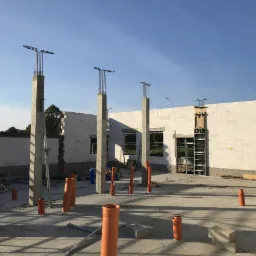 Wczesny etap budowy hali: betonowe słupy z wystającymi prętami zbrojeniowymi, ściany z bloczków betonowych, rury kanalizacyjne na posadzce, widoczne szalunki okienne.