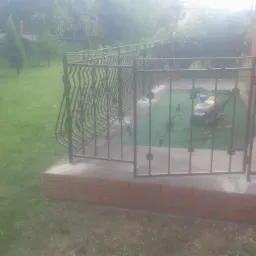 Kuta, metalowa balustrada z ozdobnymi elementami, zamontowana na ceglanym podeście przed domem, z otwartą furtką i widocznym trawnikiem w tle.