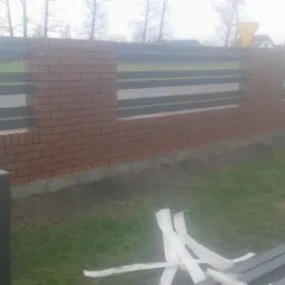 Wznoszony fragment ogrodzenia z ceglanymi słupkami i poziomymi przęsłami z metalowych profili, widoczne narzędzia i elementy montażowe na trawie.