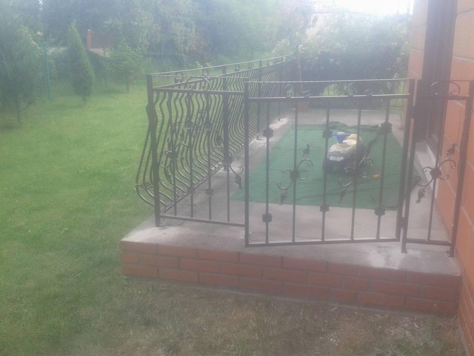 Kuta, metalowa balustrada z ozdobnymi elementami, zamontowana na ceglanym podeście przed domem, z otwartą furtką i widocznym trawnikiem w tle.