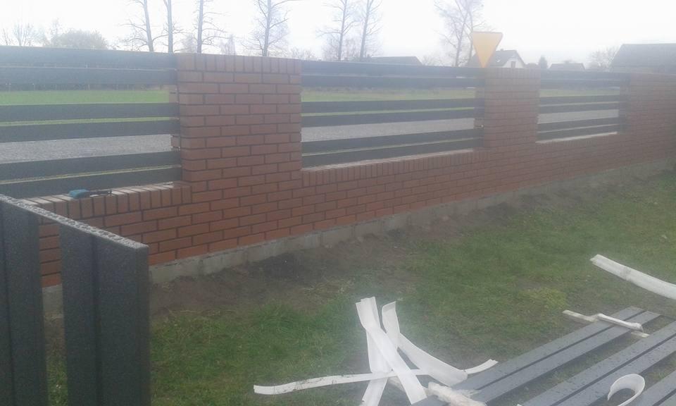 Wznoszony fragment ogrodzenia z ceglanymi słupkami i poziomymi przęsłami z metalowych profili, widoczne narzędzia i elementy montażowe na trawie.
