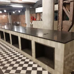 Długi, czarny blat w lokalu gastronomicznym, wsparty na podstawie wyłożonej jasnymi płytkami, z otwartymi półkami, na podłodze czarno-biała szachownica.