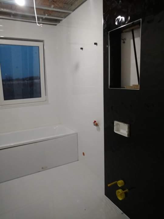 Wnętrze łazienki w trakcie prac wykończeniowych: białe ściany i podłoga, zabudowana wanna, czarna ściana z przygotowanym otworem, widoczne wystające rury i elementy instalacji.