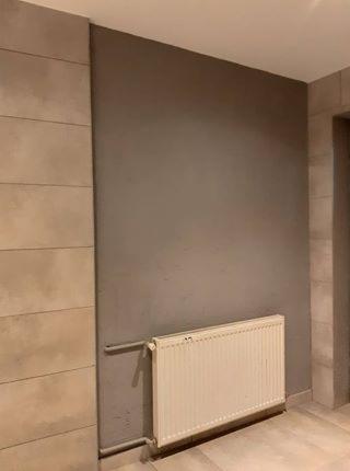 Fragment łazienki z beżowymi, poziomymi płytkami imitującymi kamień, szarą ścianą i białym grzejnikiem panelowym.