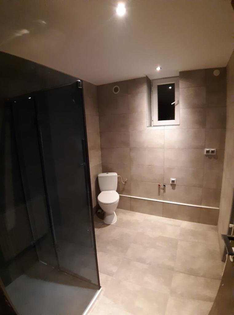 Wykończona łazienka w neutralnych barwach, z kabiną prysznicową z ciemnego szkła, wiszącą toaletą, beżowymi płytkami na ścianach i podłodze, małym oknem i minimalistycznym oświetleniem punktowym.