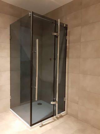 Kabina prysznicowa z ciemnego szkła z chromowanymi uchwytami i zawiasami, osadzona w narożniku wyłożonym beżowymi płytkami.
