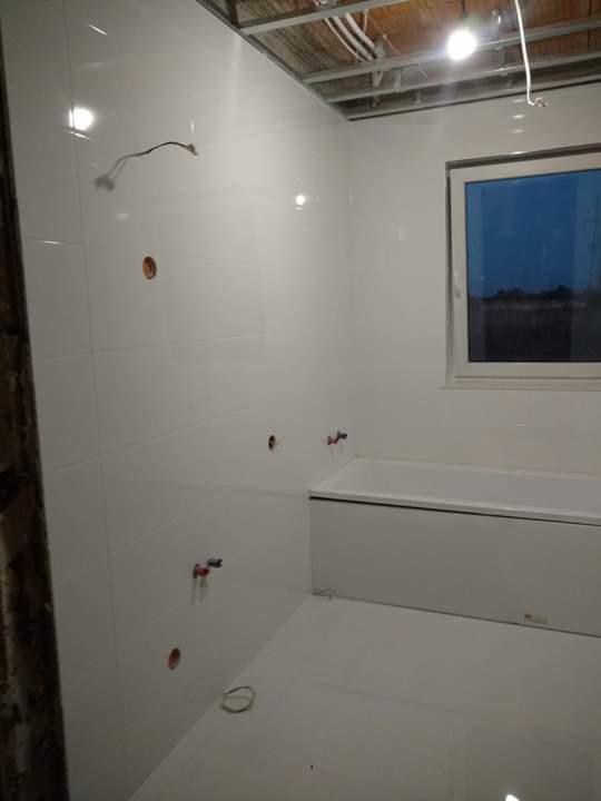 Niedokończona łazienka z białymi płytkami, widoczna wanna, instalacja wodna i sufit podwieszany w trakcie montażu.