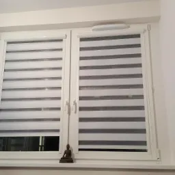 Dwuskrzydłowe okno z zamontowanymi roletami dzień-noc z białymi ramami, na parapecie mała figurka Buddy.