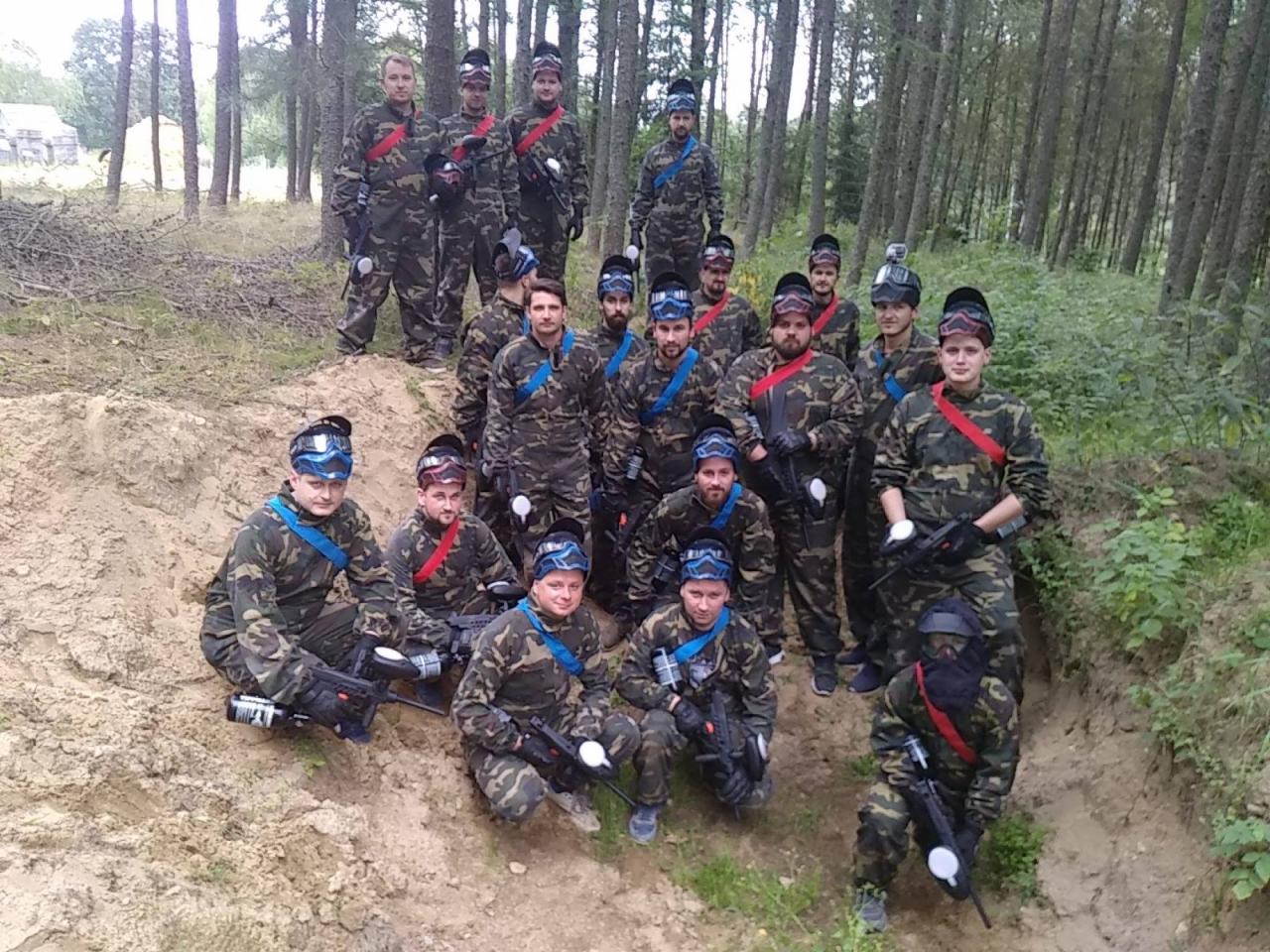 Grupa mężczyzn w strojach moro, z maskami i karabinami do paintballa, pozuje na tle lasu i piaszczystego terenu, prawdopodobnie po zakończonej grze paintballowej.