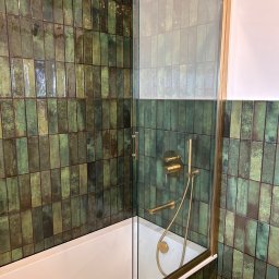 FHU ROJ - Nowoczesna łazienka z wanną i szklanym parawanem w złotej ramie. Ściany wyłożone zielonymi, podłużnymi płytkami. Złota armatura.