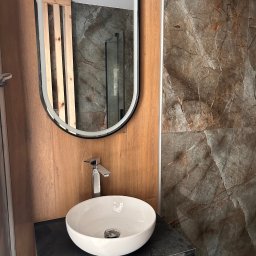 FHU ROJ - Nowoczesna łazienka z okrągłą umywalką, lustrem w czarnej ramie i płytkami imitującymi marmur. Widoczne drewniane panele i szklana kabina prysznicowa.