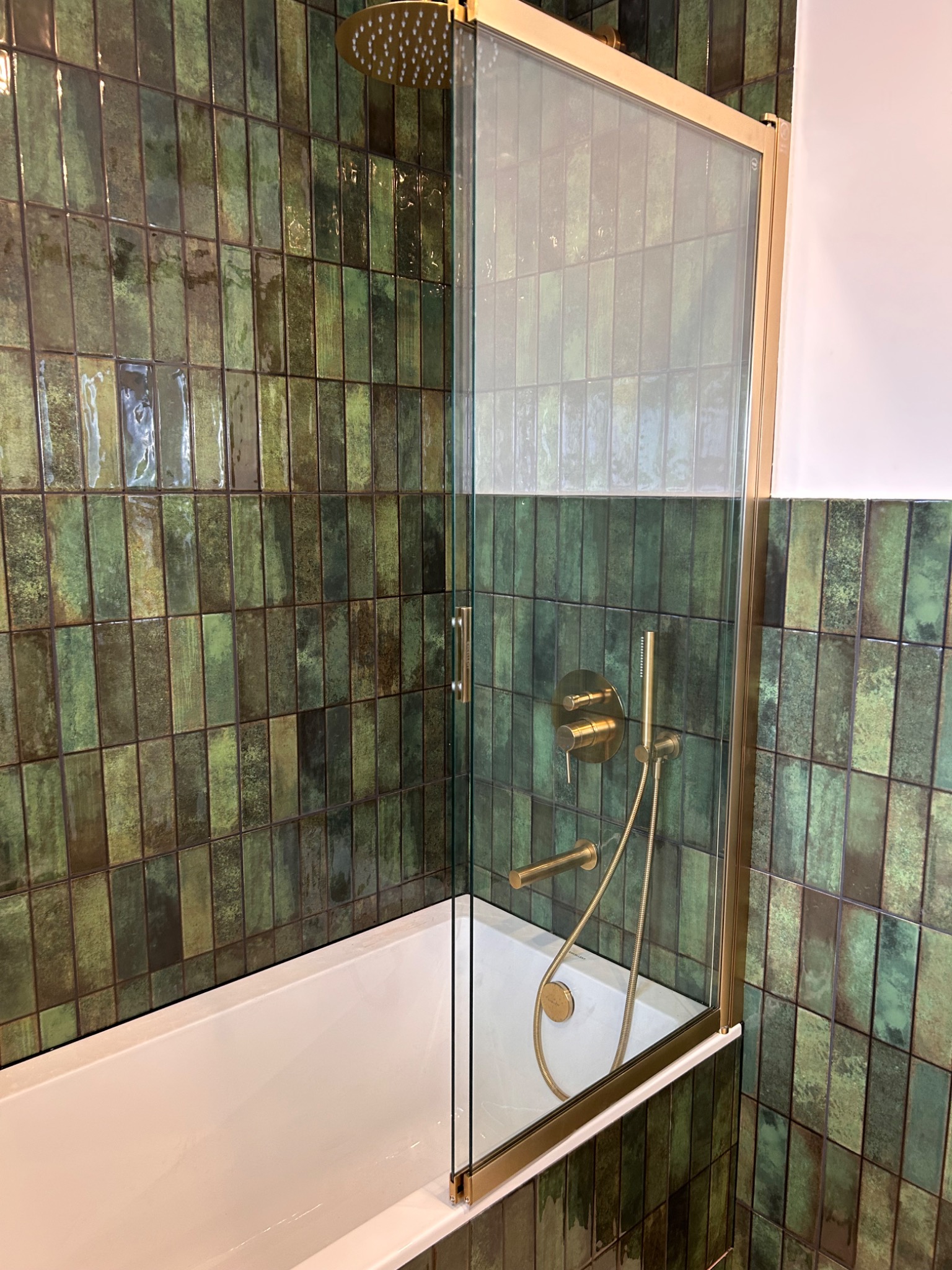 Nowoczesna łazienka z wanną i szklanym parawanem w złotej ramie. Ściany wyłożone zielonymi, podłużnymi płytkami. Złota armatura.