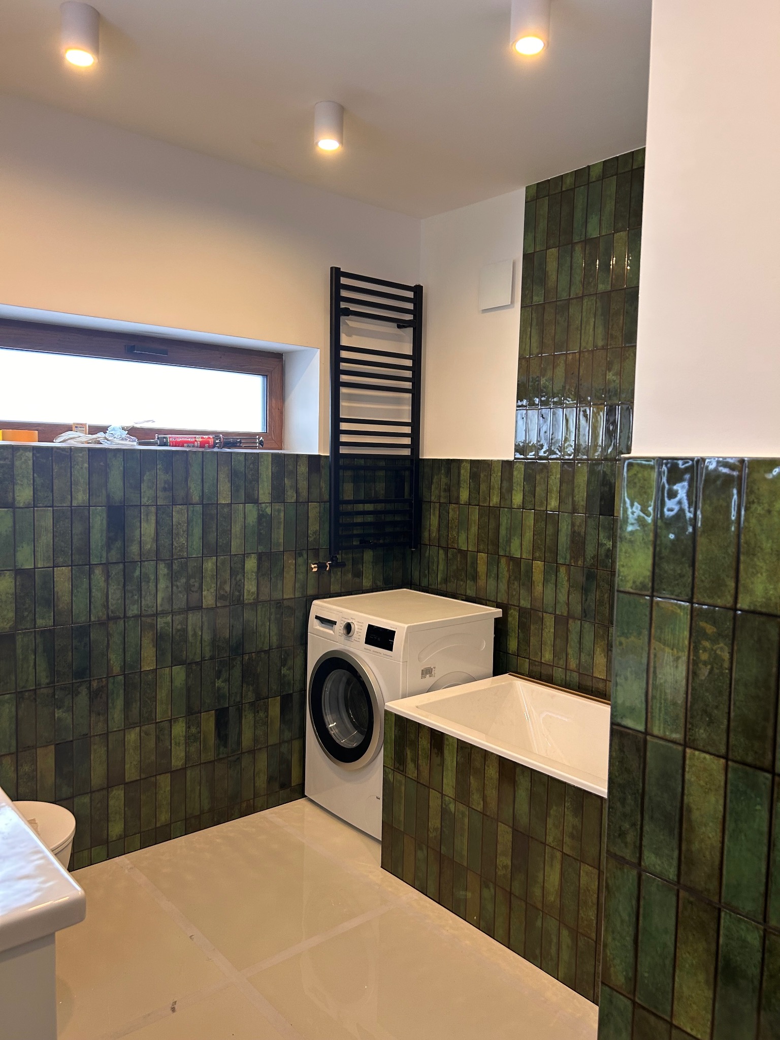 Łazienka po remoncie z zielonymi, błyszczącymi płytkami ułożonymi pionowo, białą wanną i pralką. Widoczne okno i grzejnik drabinkowy. Jasna podłoga.