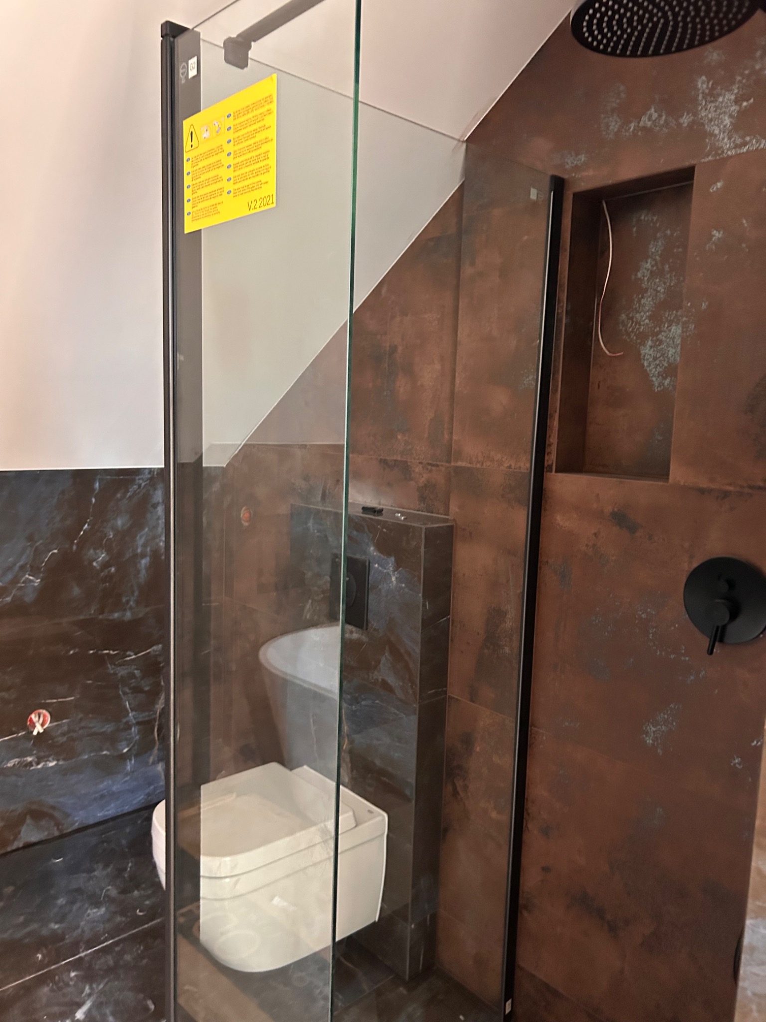 Nowoczesna kabina prysznicowa z czarnymi detalami i oryginalnymi płytkami w odcieniach brązu i granatu. Widoczna bateria i półka w ścianie.