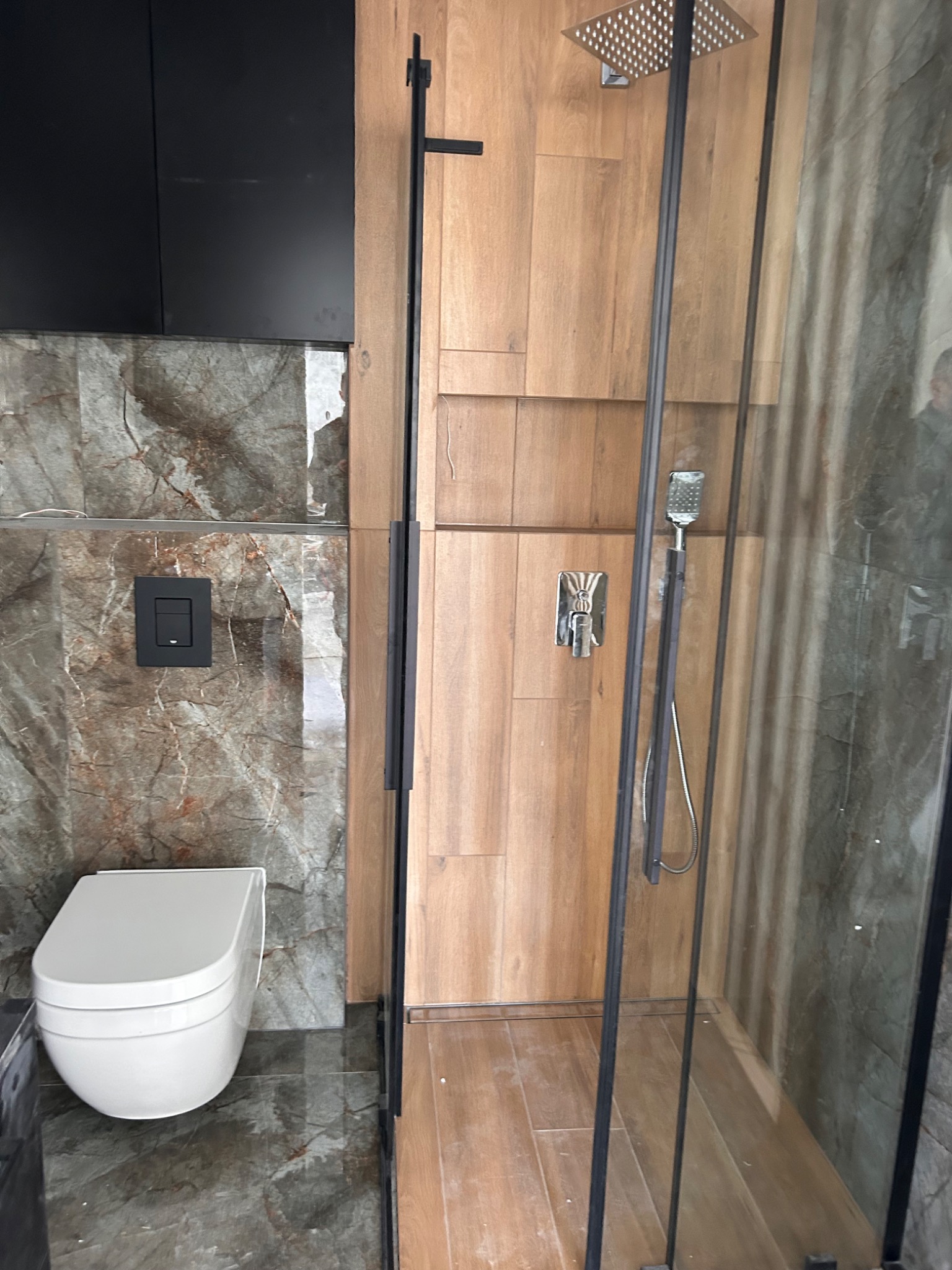 Nowoczesna łazienka z kabiną prysznicową typu walk-in, drewnopodobnymi płytkami i wiszącą toaletą. Minimalistyczny design z czarnymi akcentami.