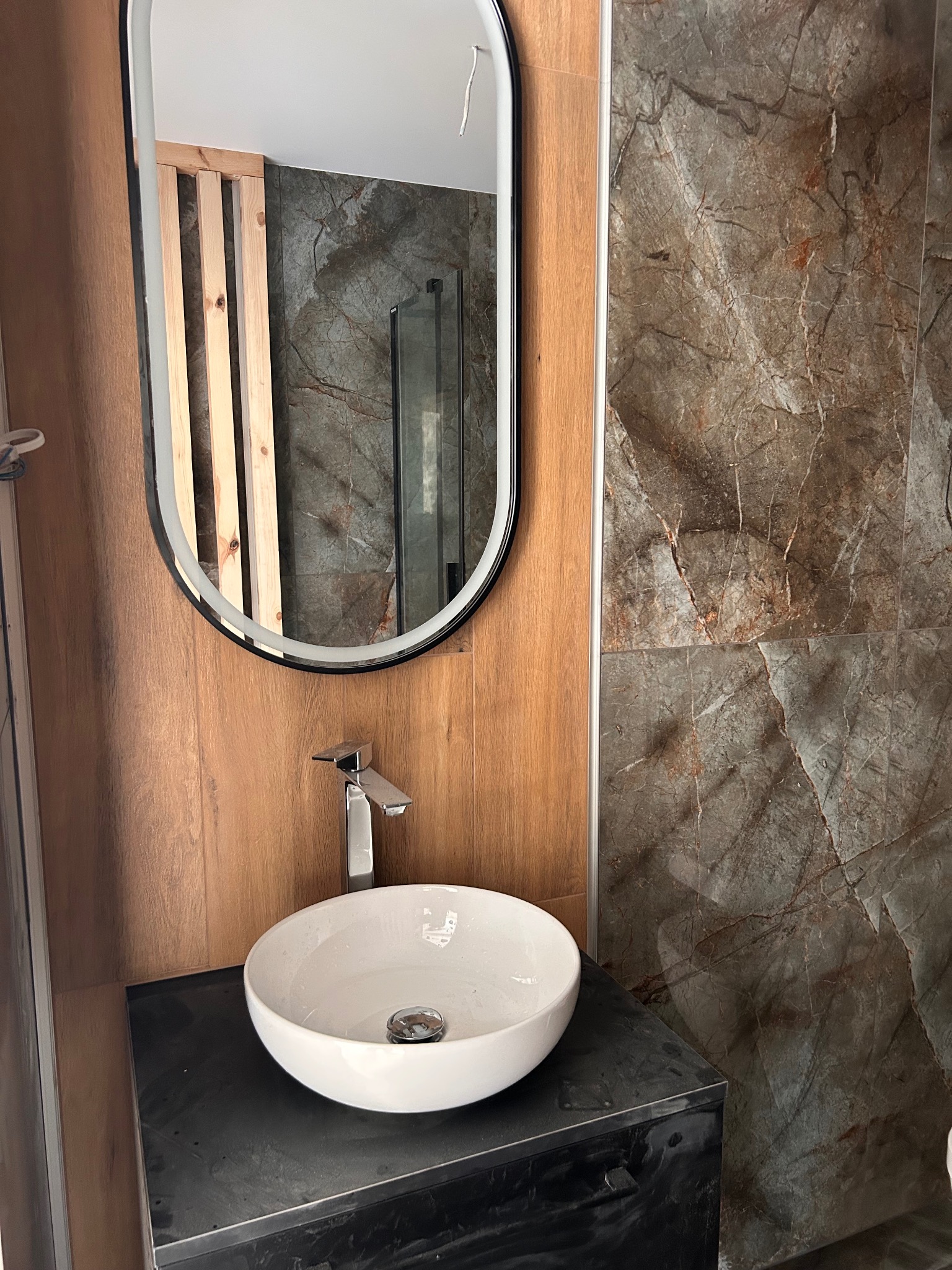 Nowoczesna łazienka z okrągłą umywalką, lustrem w czarnej ramie i płytkami imitującymi marmur. Widoczne drewniane panele i szklana kabina prysznicowa.