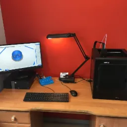 Biurko z komputerem wyświetlającym model 3D i drukarką 3D Zortrax M200 Plus, oświetlone lampką biurkową, na tle czerwonej ściany.