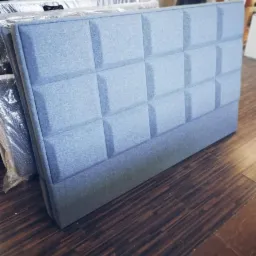 Błękitny, tapicerowany zagłówek z geometrycznym wzorem kwadratów, leżący na podłodze z paneli.