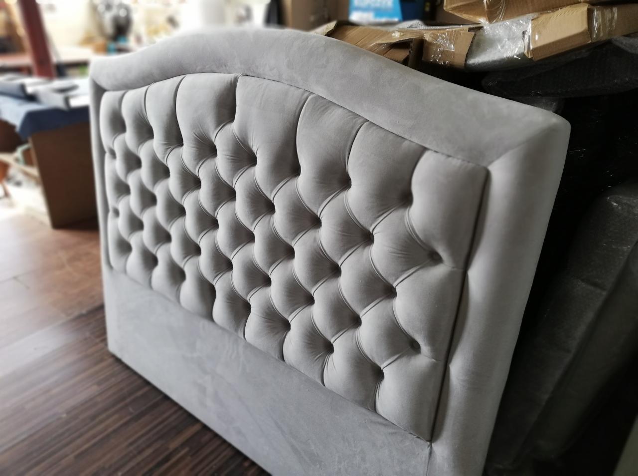 Szary, tapicerowany zagłówek łóżka z pikowaniem w romby, widoczny w magazynie lub warsztacie, z kartonami w tle.