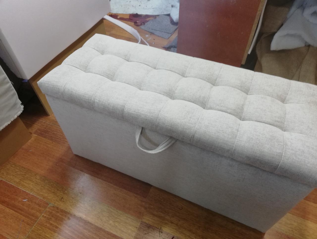 Tapicerowany puf z pikowanym siedziskiem w jasnym odcieniu szarości, z uchwytem z boku, na drewnianej podłodze, w tle widoczne elementy warsztatu.