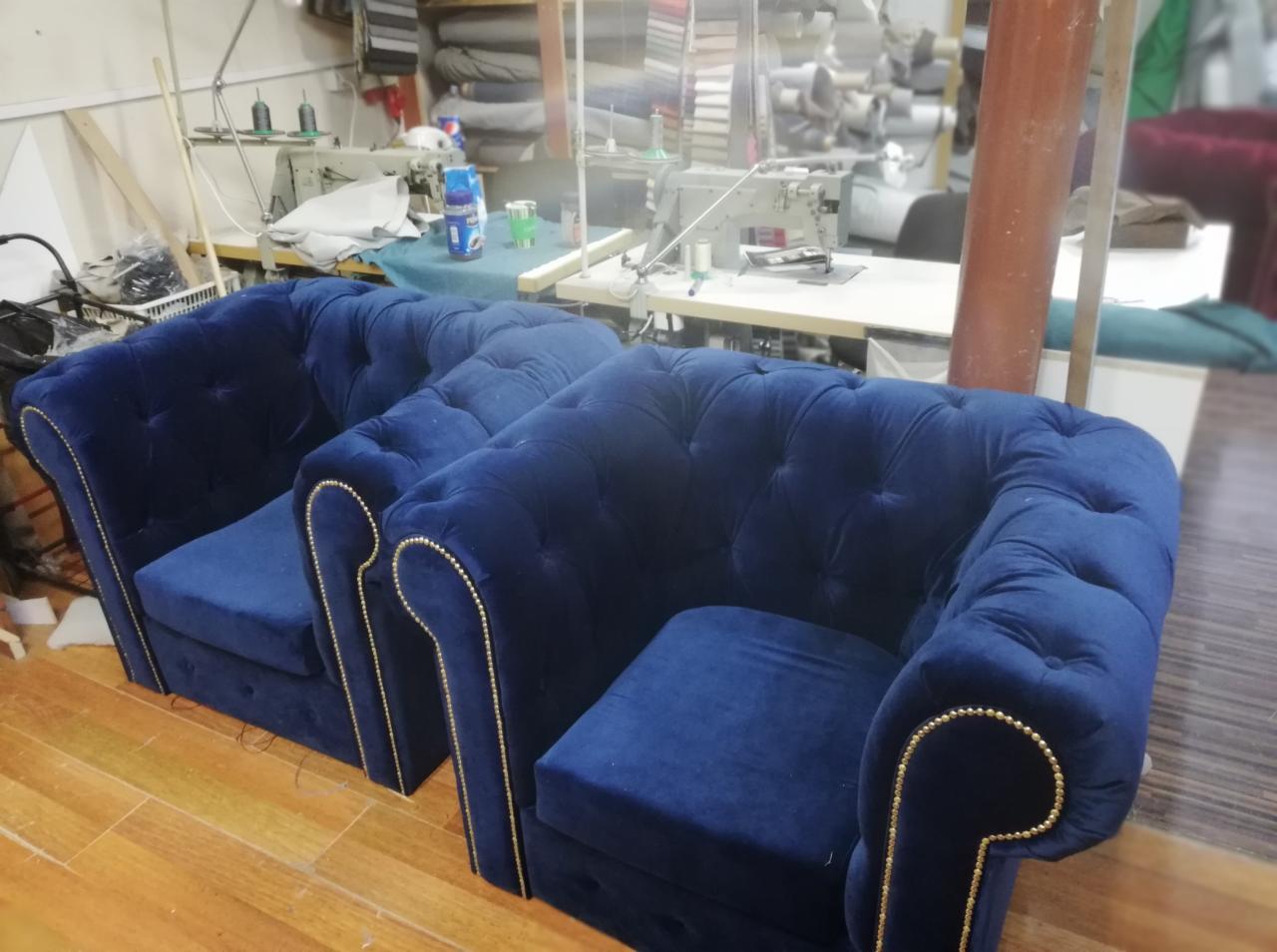 Dwa granatowe fotele klubowe typu Chesterfield z ozdobnymi pinezkami, wykonane w Pruszkowie, widoczne na tle warsztatu tapicerskiego z maszynami do szycia.