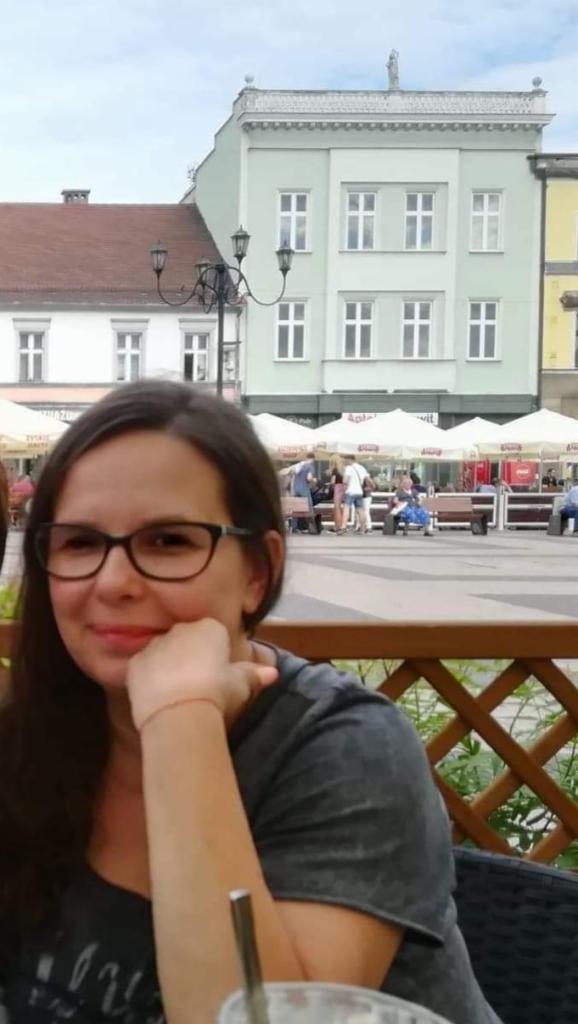 Kobieta w okularach siedzi w kawiarni na zewnątrz, na tle plac miejski z zabytkowymi budynkami i parasolami.