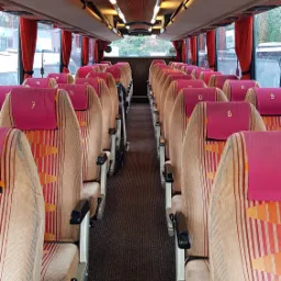 Wnętrze autobusu z rzędami foteli pasażerskich w beżowo-pomarańczowe pasy, z różowymi zagłówkami oznaczonymi numerami, widok z przodu w kierunku tyłu pojazdu.