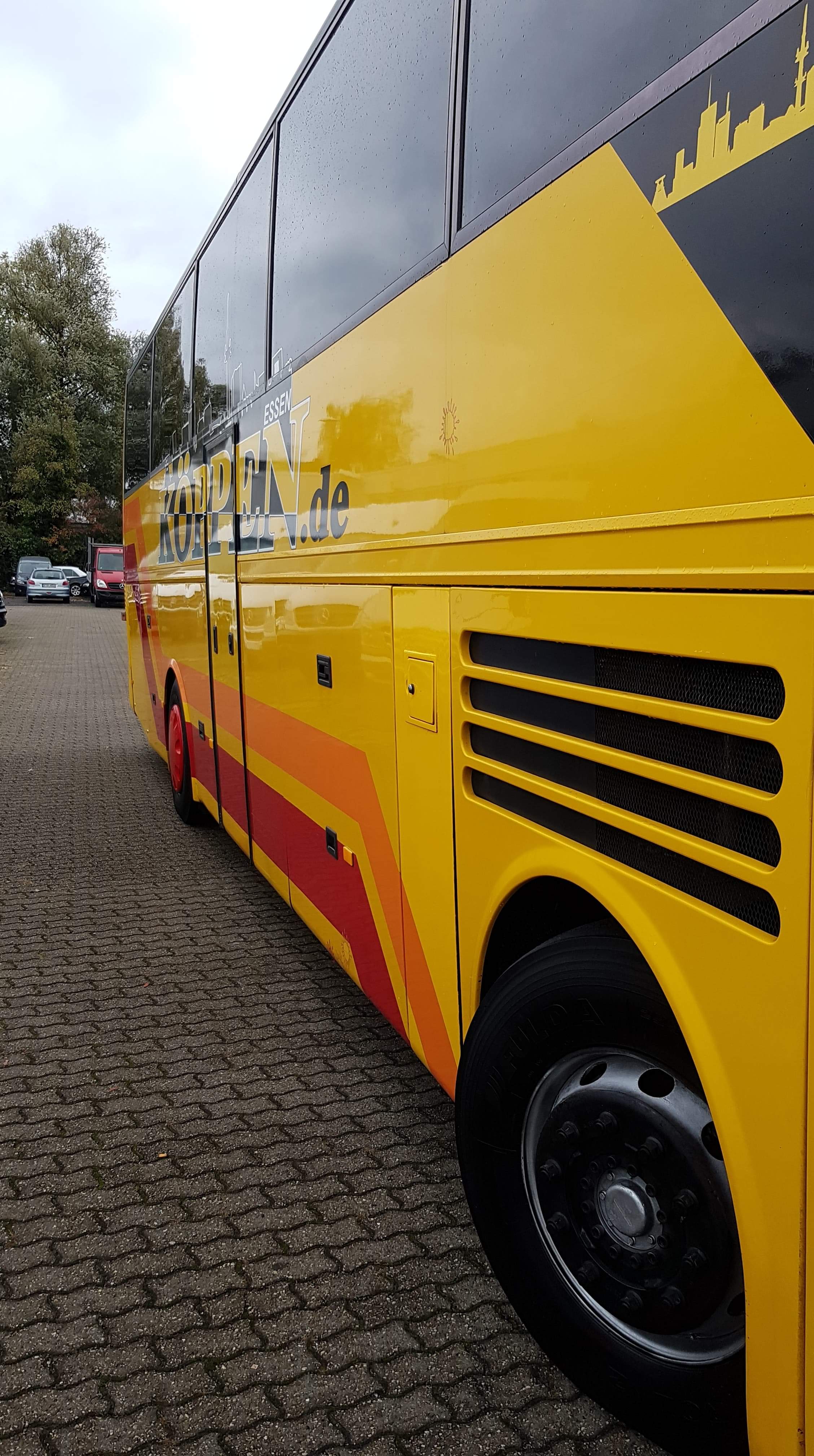 Żółty autokar z czerwonymi i pomarańczowymi pasami, logo firmy Köppen.de i sylwetką miasta Essen na tle zachmurzonego nieba, zaparkowany na kostce brukowej.