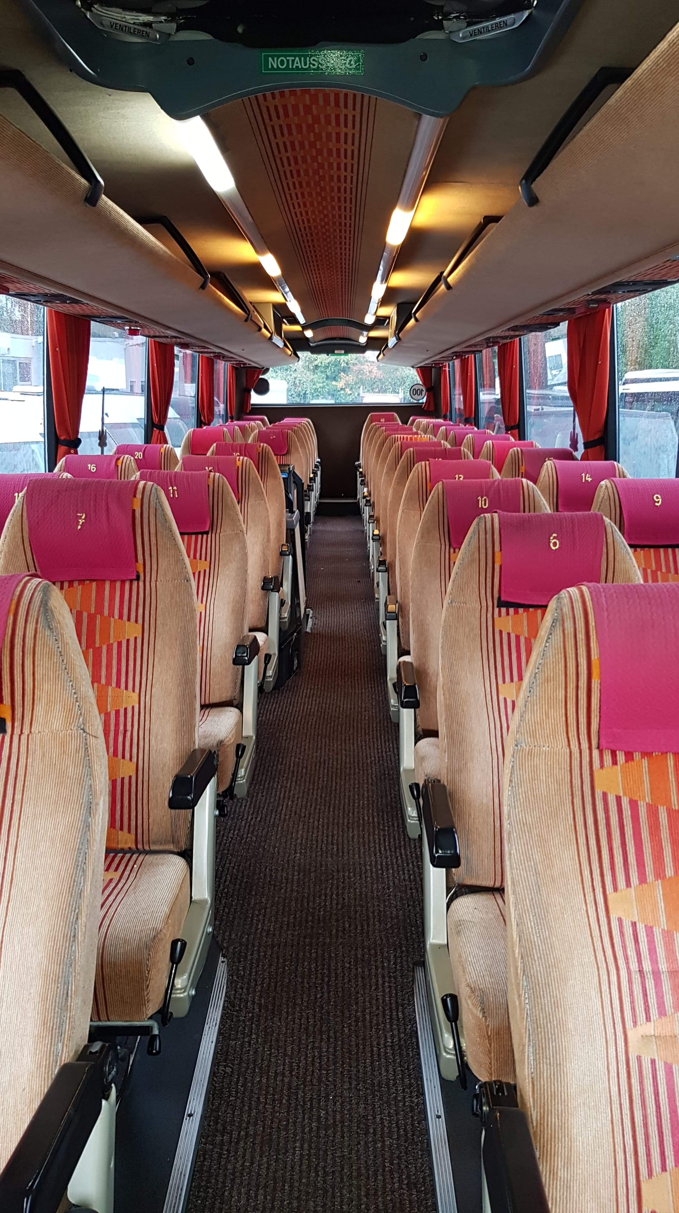 Wnętrze autobusu z rzędami foteli pasażerskich w beżowo-pomarańczowe pasy, z różowymi zagłówkami oznaczonymi numerami, widok z przodu w kierunku tyłu pojazdu.