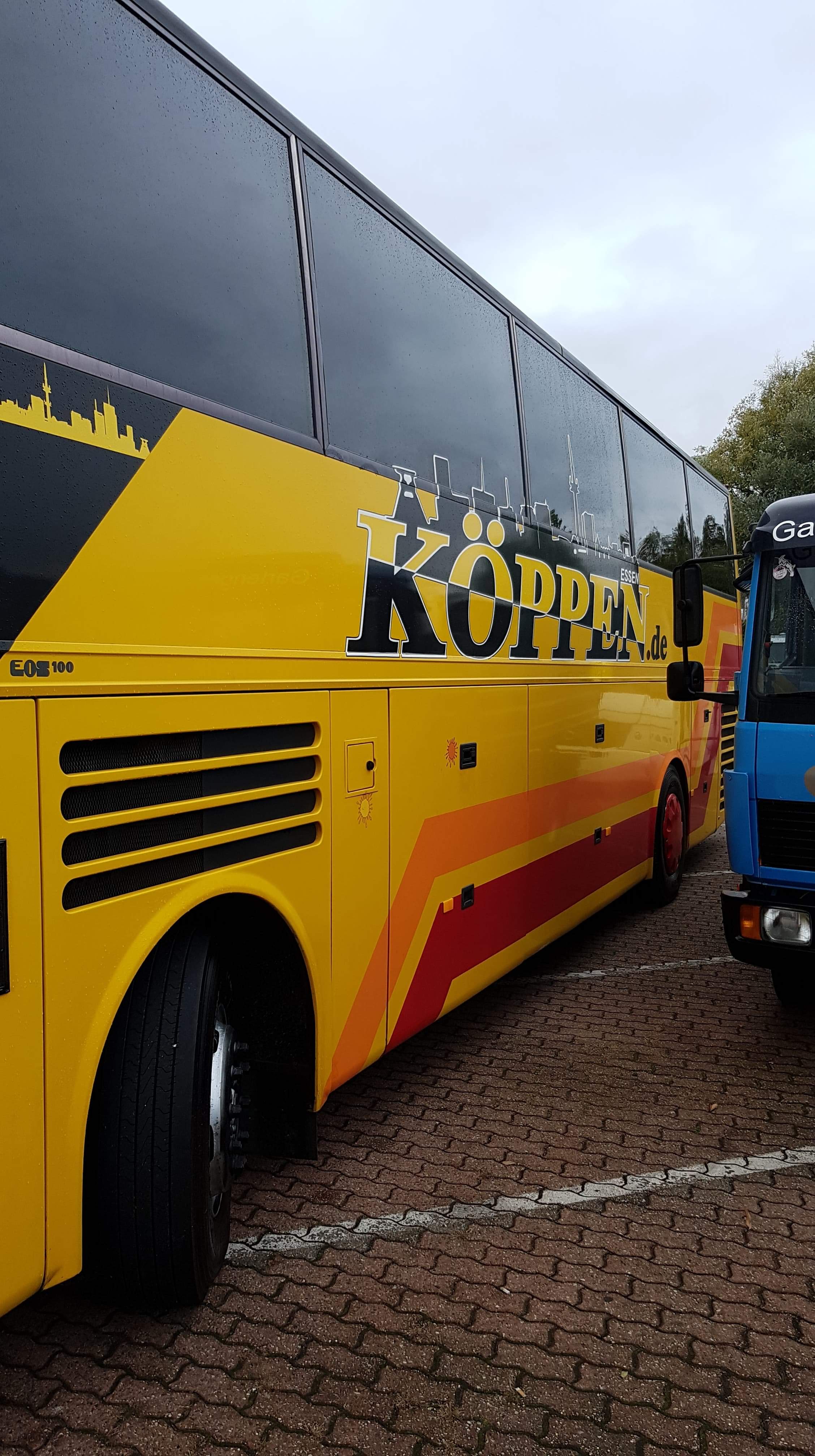 Żółty autobus z logo firmy Köppen i sylwetką miasta Essen, zaparkowany na brukowanym placu, obok niebieskiej ciężarówki.