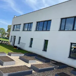 Kwadratowe, podwyższone rabaty wypełnione ziemią na żwirowym podłożu przed nowoczesnym budynkiem z ciemnymi oknami.