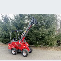 Czerwona mini ładowarka teleskopowa z podniesionymi widłami na tle zielonych drzew i betonowej nawierzchni.