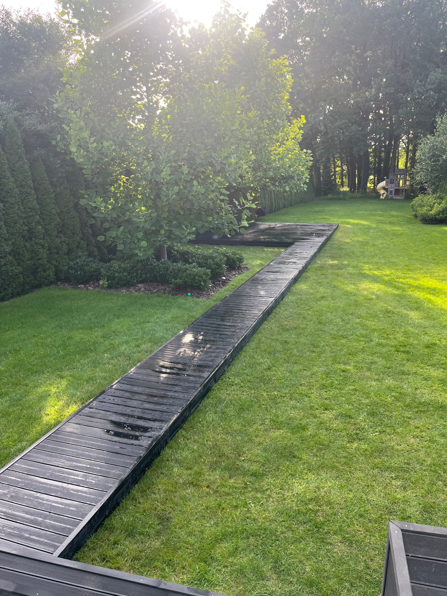 Drewniana ścieżka w ogrodzie w Otrębusach, prowadząca przez trawnik w kierunku drzew i placu zabaw, wykonana z ciemnego kompozytu, lekko wilgotna, w słoneczny dzień.