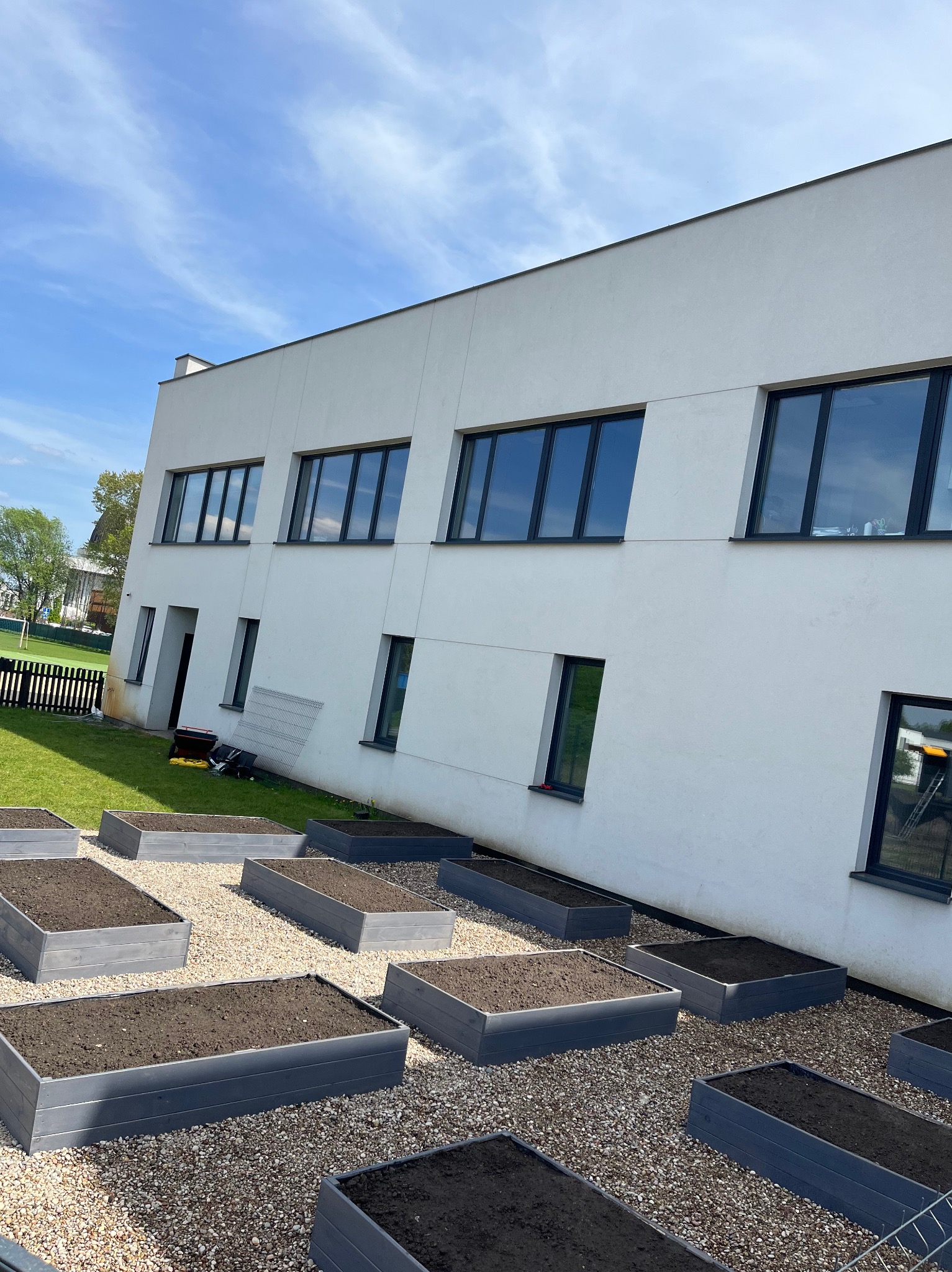 Kwadratowe, podwyższone rabaty wypełnione ziemią na żwirowym podłożu przed nowoczesnym budynkiem z ciemnymi oknami.