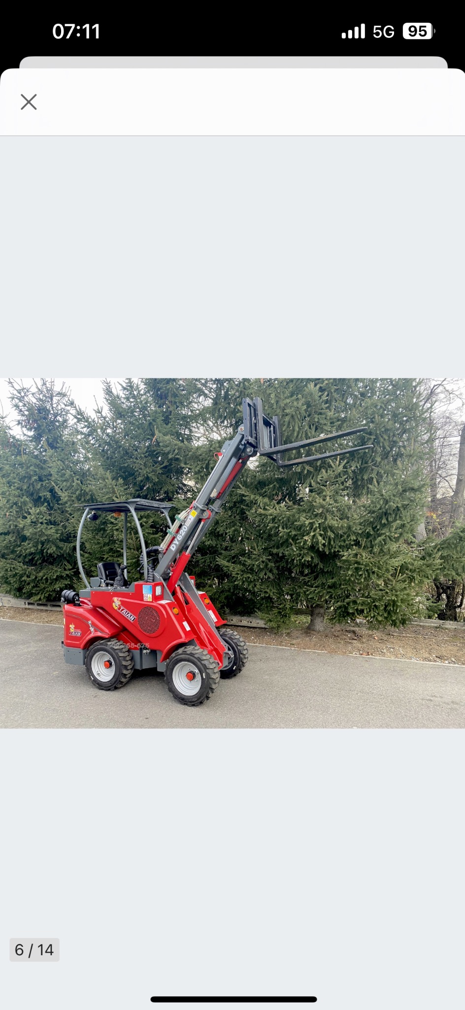 Czerwona mini ładowarka teleskopowa z podniesionymi widłami na tle zielonych drzew i betonowej nawierzchni.