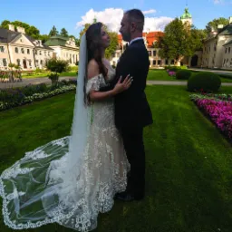 Para młoda w objęciach na tle klasycystycznego pałacu z wieżą, otoczona zielonym trawnikiem i rabatami kwiatowymi w odcieniach fioletu i różu. Suknia panny młodej z koronkowymi zdobieniami i długim...