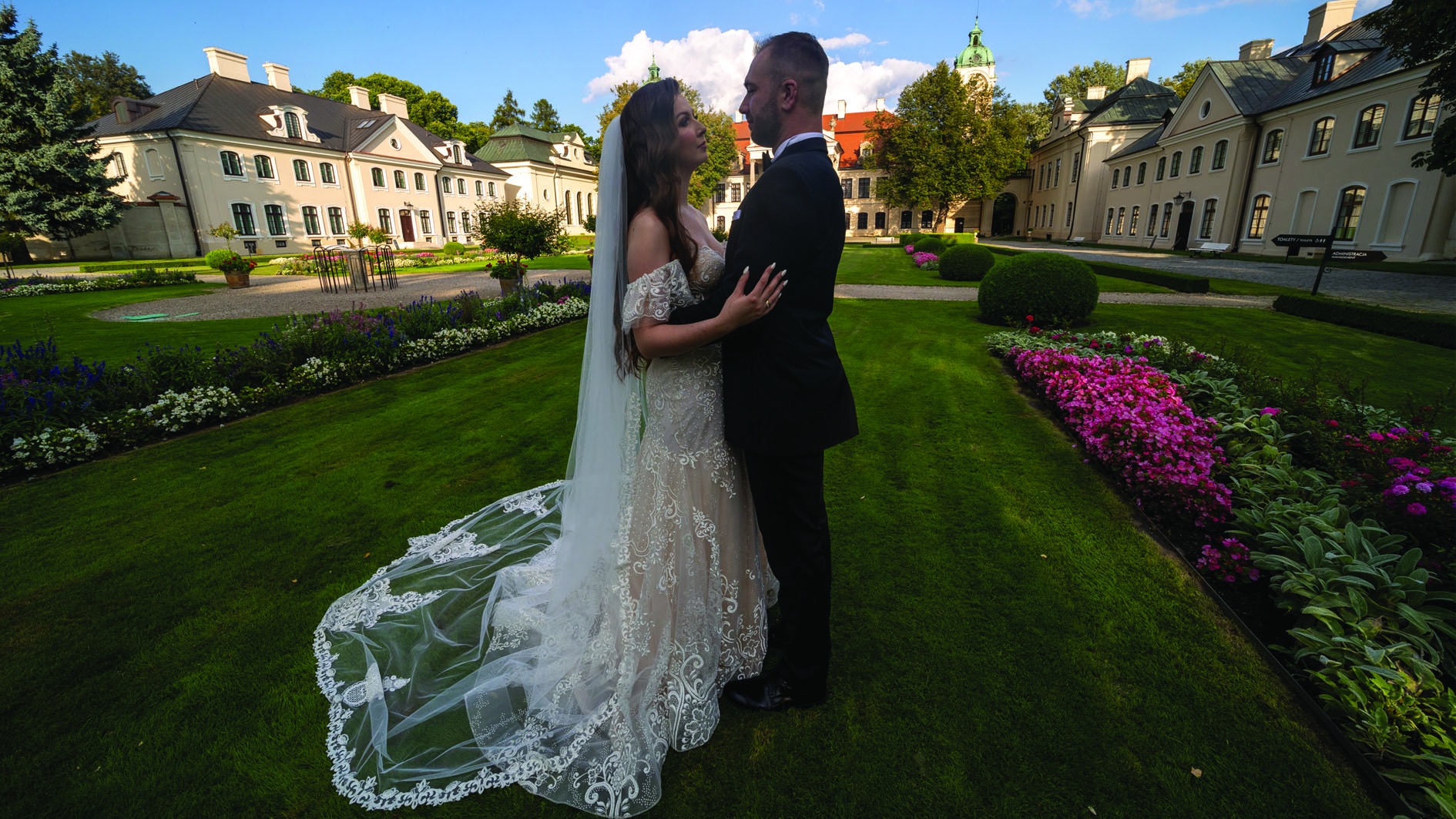 Para młoda w objęciach na tle klasycystycznego pałacu z wieżą, otoczona zielonym trawnikiem i rabatami kwiatowymi w odcieniach fioletu i różu. Suknia panny młodej z koronkowymi zdobieniami i długim...