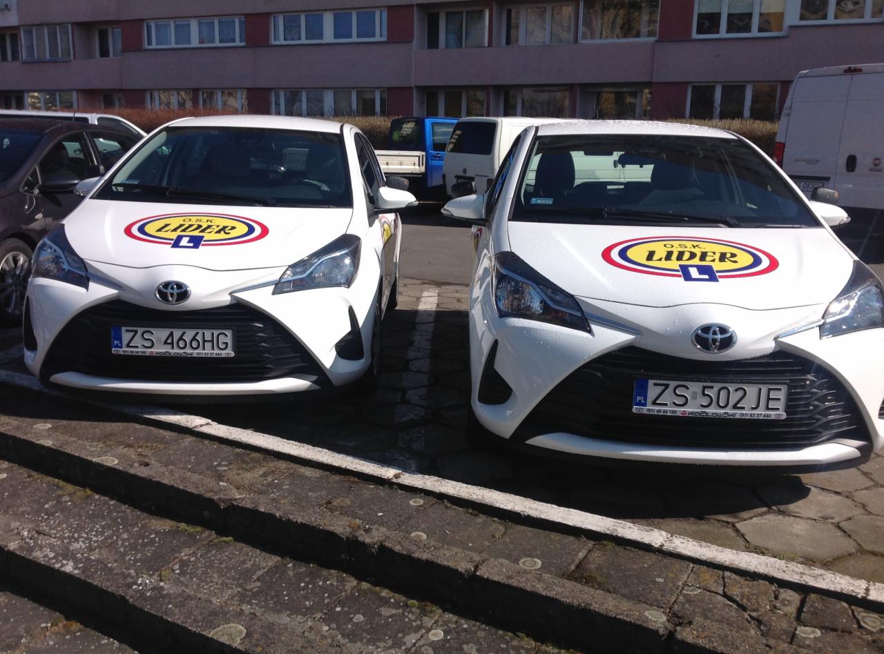 Dwa białe samochody Toyota Yaris z logo O.S.K. Lider na masce, zaparkowane obok siebie na brukowanym parkingu przed budynkiem.