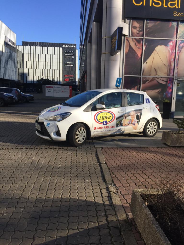 Biały samochód z naklejkami 'L' i logiem ośrodka szkolenia kierowców zaparkowany na tle nowoczesnego budynku galerii handlowej w słoneczny dzień.