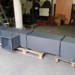 Szary, prostokątny kanał wentylacyjny z bocznym otworem, ułożony na drewnianych paletach w warsztacie.