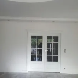 Jasne, białe dwuskrzydłowe drzwi wewnętrzne z szybami, zamontowane w Jeleniej Górze, widoczne na tle białych ścian i sufitu z oświetleniem punktowym.