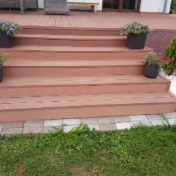 Schody z desek kompozytowych prowadzące na taras, ozdobione donicami z kwiatami i białymi kamieniami dekoracyjnymi, widoczne trawnik i elementy architektury domu.