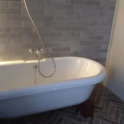 Biała wanna wolnostojąca z chromowaną baterią, ustawiona na drewnianych podkładkach, na podłodze ułożonej w jodełkę z płytek imitujących drewno. Ściana wyłożona szarą cegłą z podświetleniem LED...