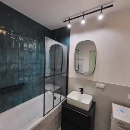 Nowoczesna łazienka z ciemnozielonymi, połyskującymi płytkami, białą wanną z czarną szybą prysznicową, umywalką nablatową na czarnej szafce i owalnym lustrem z czarną ramą.