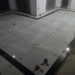 Układanie jasnych płytek podłogowych z ciemną bordiurą, widoczne krzyżyki dystansowe, miarka i obcinak do płytek w rogu pomieszczenia w trakcie prac wykończeniowych.