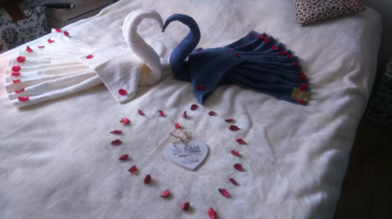 Dwa łabędzie uformowane z ręczników, biały i granatowy, ustawione na łóżku z pościelą w kolorze ecru, udekorowane płatkami czerwonych róż tworzącymi kształt serca wokół drewnianego serca z napisem.