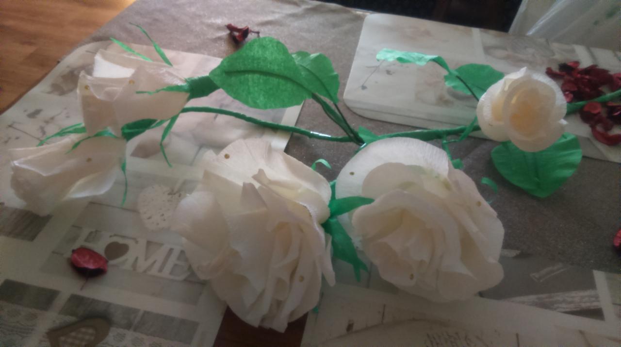 Ręcznie robione róże z białej bibuły z zielonymi łodygami i liśćmi, leżące na wzorzystym obrusie.