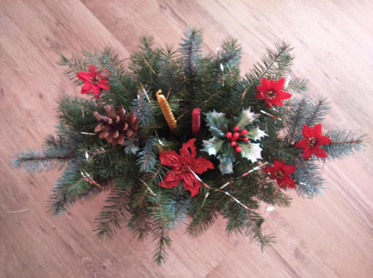 Dekoracja świąteczna ze świerkowych gałązek, czerwonych kwiatów, szyszki, ostrokrzewu i dwóch świec, ułożona na drewnianej podłodze.