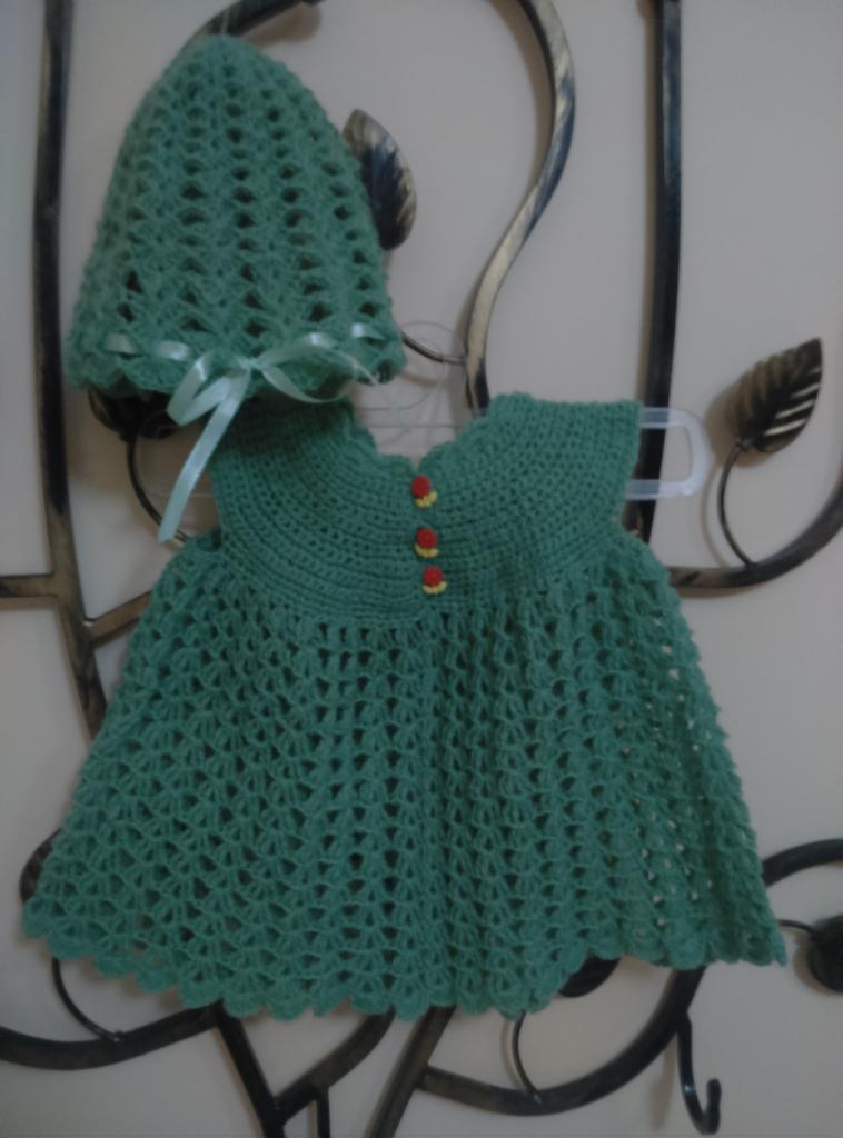 Zielona, ażurowa sukienka dla dziewczynki z dopasowanym kapelusikiem, ozdobionym wstążką, zawieszona na ozdobnym, metalowym wieszaku.