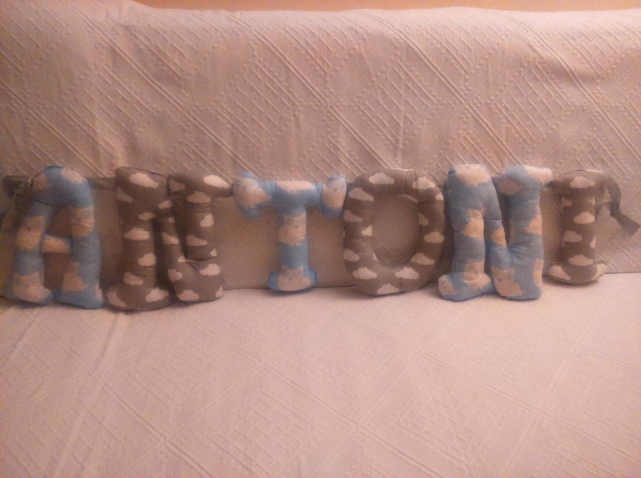 Poduszki dekoracyjne w kształcie liter układające się w imię 'ANTONIO', wykonane z materiałów w odcieniach szarości i błękitu, ozdobione wzorem chmurek i sów, leżące na białej pikowanej narzucie.